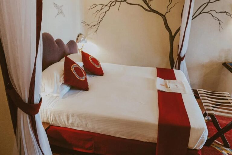 Ein großes Bett mit roten Kissen zwischen zwei weißen Vorhängen, vor einem an die Wand gemalten Baum