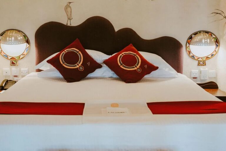 Zwei bestickte rote Kissen auf einem großen Bett, unter einem an die Wand gemalten Reiher und zwischen zwei bunten Wandleuchten