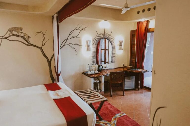 Ein Zimmer mit Gepäckablage, Schreibtisch, Wasserkocher und kleinem Kühlschrank unter einem Deckenventilator