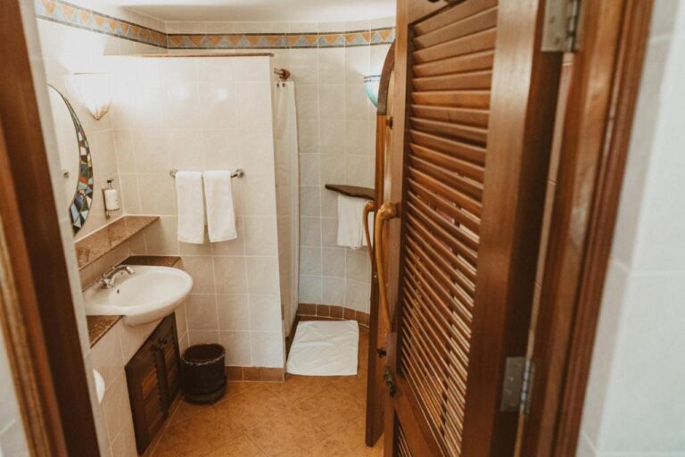 Ein Bad, gesehen von seiner Holztür mit Lamellen, mit Waschbecken und Dusche mit Vorhang