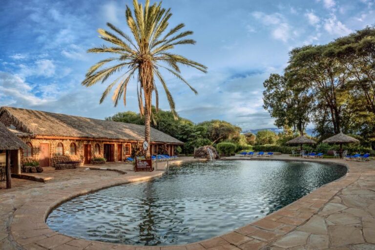 Der Pool der Amboseli Sopa Lodge mit einer Palme, Strohschirmen und einem Steingebäude