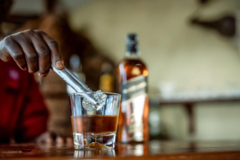 Eine Hand legt einen Eiswürfel in ein Glas Whisky, auf der Theke der Bar