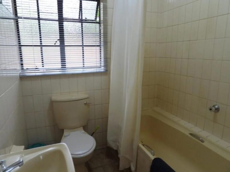 Ein gefliestes Bad mit Badewanne und Duschvorhang, WC und Waschbecken, unter einem Fenster mit Rollo