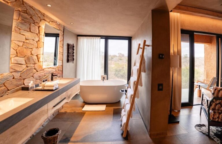 Das Bad einer Suite mit einer Badewanne vor dem Fenster und einem Doppelwaschbecken an einer Steinwand