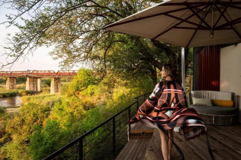 Eine Frau im Bademantel auf einer Holzterrasse unter einem Sonnenschirm, mit Blick auf Brücke und Fluss