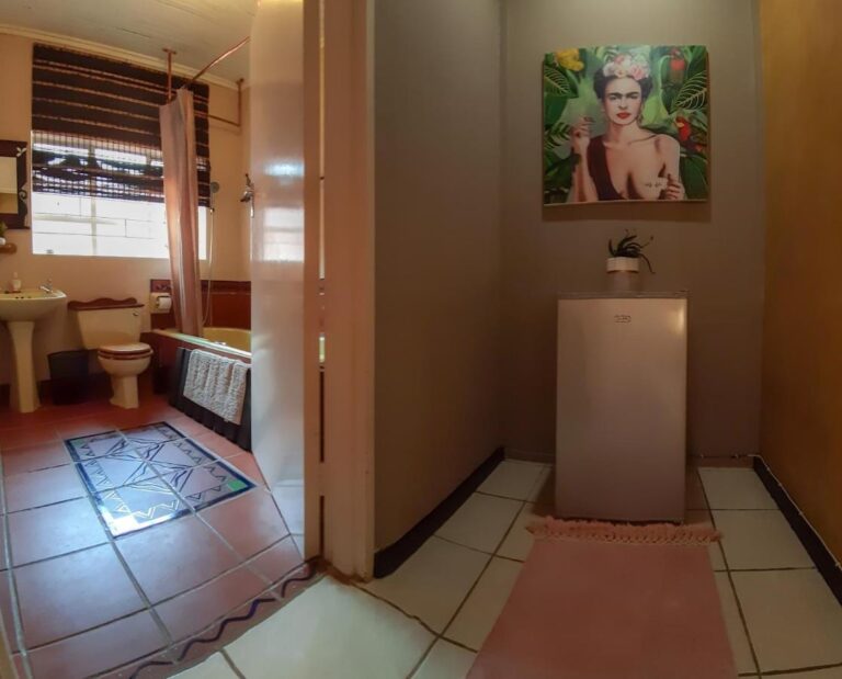 Ein Bad mit Badewanne, Waschbecken und WC, von einem Flur aus gesehen, in dem ein kleiner Kühlschrank unter einem Frauenporträt steht