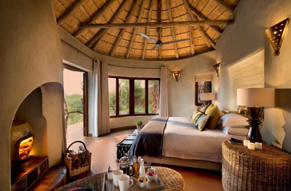Eine Suite der Madikwe Safari Lodge unter einem Strohdach, mit großem Bett und brennendem Kamin