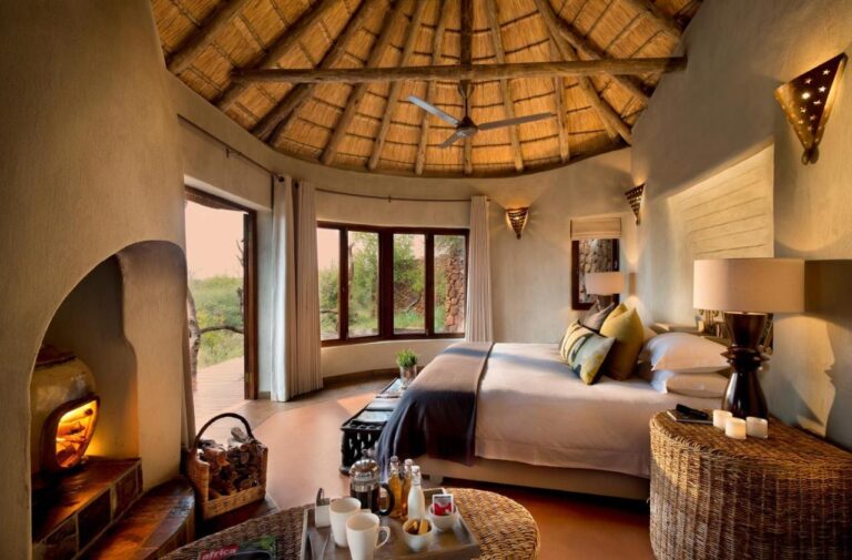 Eine Suite der Madikwe Safari Lodge unter einem Strohdach, mit großem Bett und brennendem Kamin