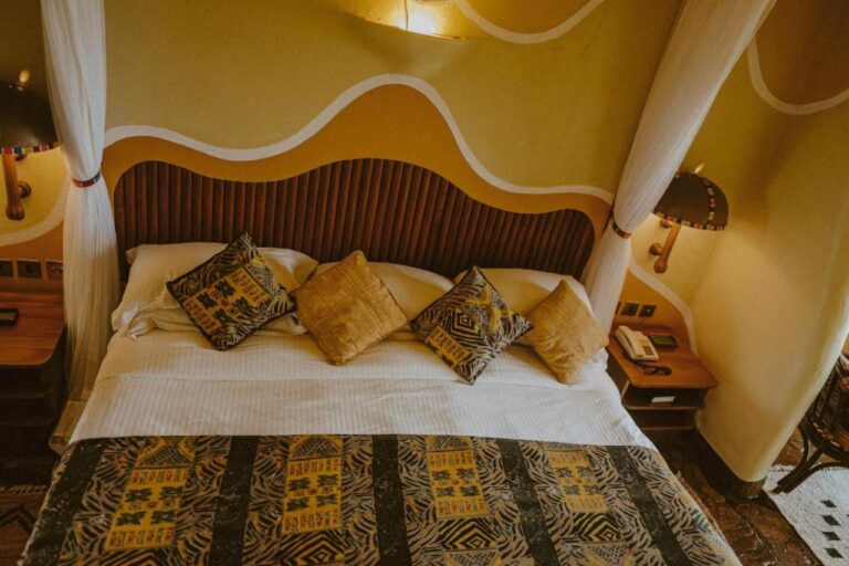 Ein großes Bett mit Kissen in afrikanischen Mustern, unter einem Kopfteil aus Holzlatten und wellenförmig bemalten Wänden