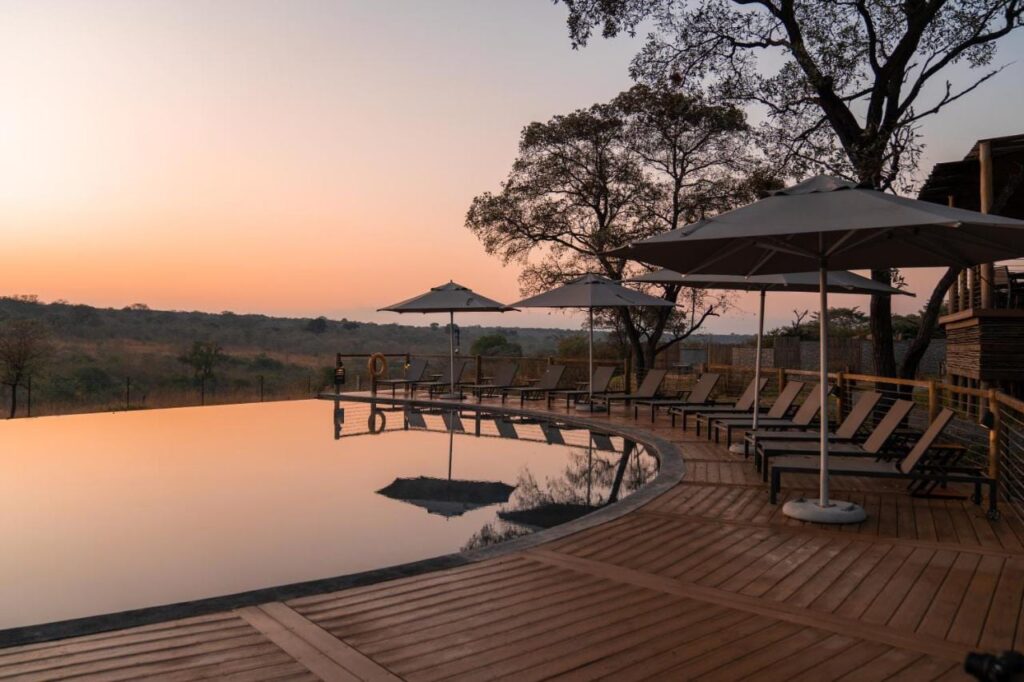 Der Infinity-Pool der Mdluli Safari Lodge unter orangefarbenem Himmel, gesäumt von Liegestühlen und Sonnenschirmen