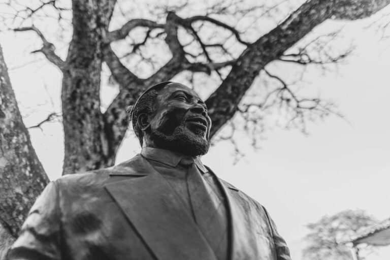 Die Bronzestatue eines bärtigen, lächelnden Mannes im Anzug unter einem Baum, in Schwarzweiß