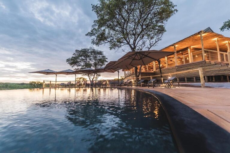 Der Infinity-Pool der Mdluli Safari Lodge und das beleuchtete Hauptgebäude unter bewölktem Himmel