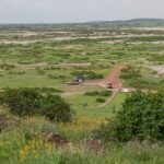 Blick von Observation Hill auf eine grüne, salzgefleckte Ebene, eine rote Piste, drei geparkte 4x4 neben einem kleinen ockerfarbenen Gebäude, niedrige Hügel in der Ferne
