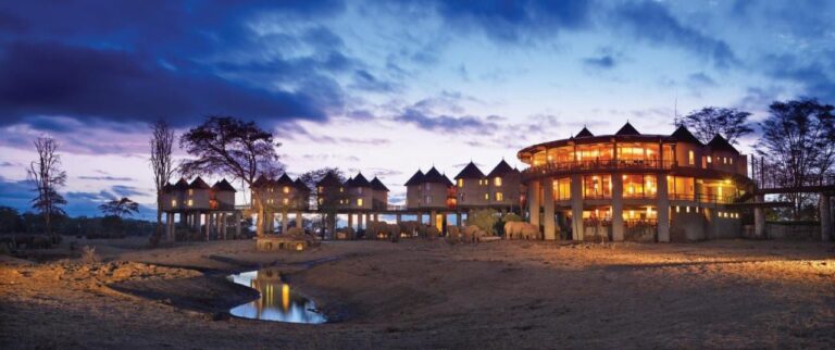 Die beleuchtete Lodge bei Einbruch der Nacht, mit Elefanten am Fuß der Türme und einer Wasserstelle im Vordergrund