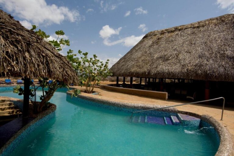 Der Pool mit blauem Mosaik, zwischen einer Strohhütte und einem großen Gebäude mit Strohdach