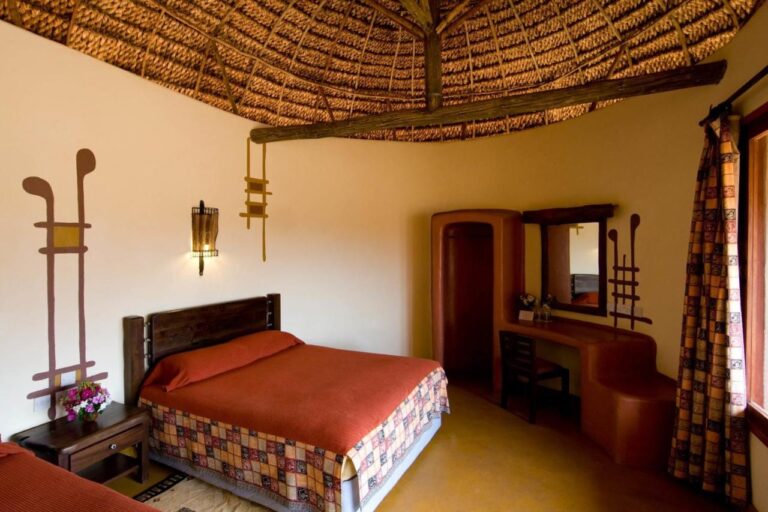 Ein Zimmer mit großem, orange bezogenem Bett unter einer Strohdecke, mit einem ockerfarbenen, abgerundeten Frisiertisch