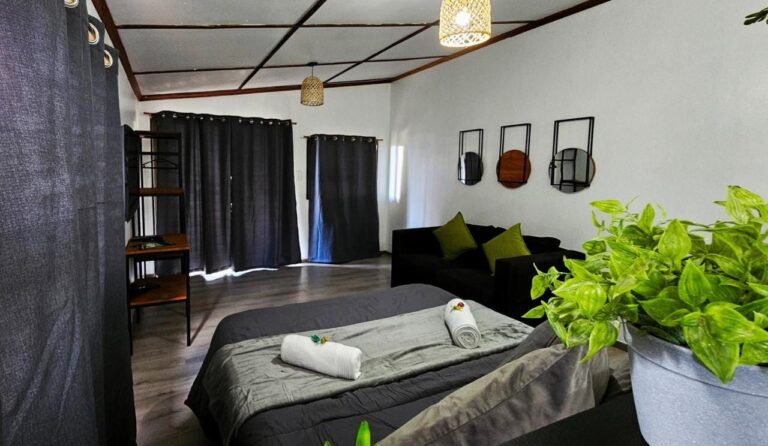 Ein großes graues Bett mit gerollten Handtüchern, ein schwarzes Sofa und dunkle Vorhänge, in einem Studio mit Holzkassettendecke