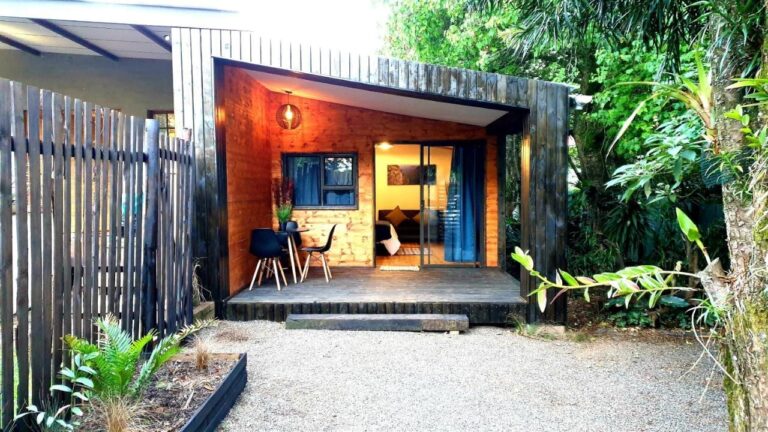 Ein Holzchalet mit überdachter Terrasse, Tisch und zwei schwarzen Stühlen, vor einem Kieshof und Pflanzen