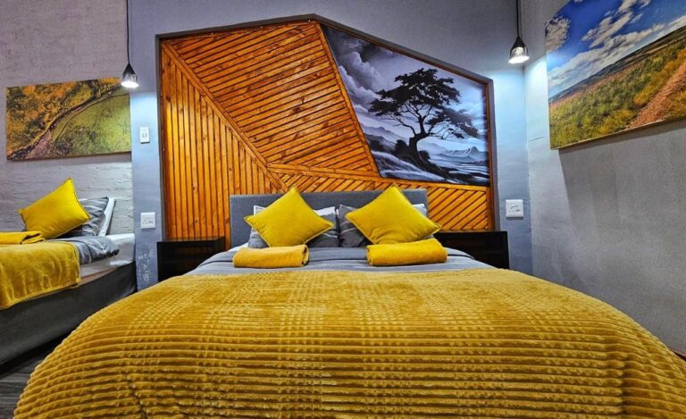 Ein großes Bett mit gelber Decke vor einem Kopfteil aus Holzlatten und einem Schwarz-Weiß-Gemälde eines Baumes, neben einem zweiten Bett
