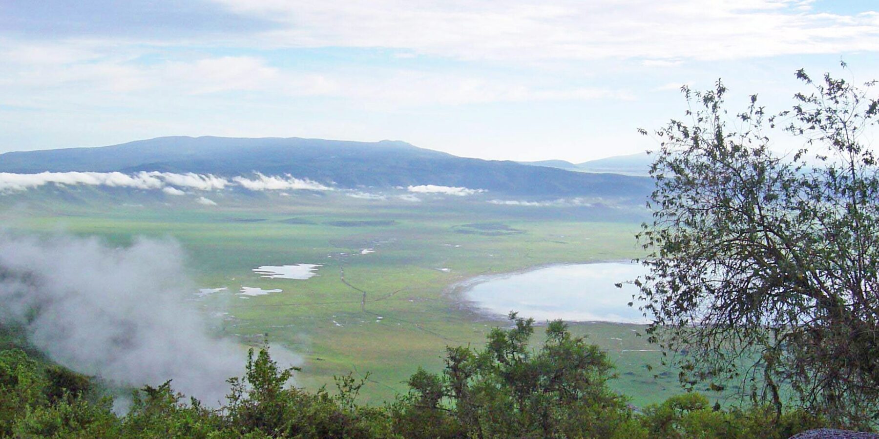 Der grüne Kraterboden vom Rand aus gesehen, mit dem Magadi-See und Wolken an den Kraterwänden